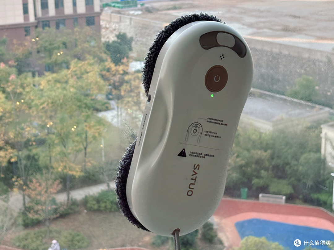 洒拖T30擦窗机器人工作特写，双圆盘设计清晰可见，正吸附在明亮的玻璃上。