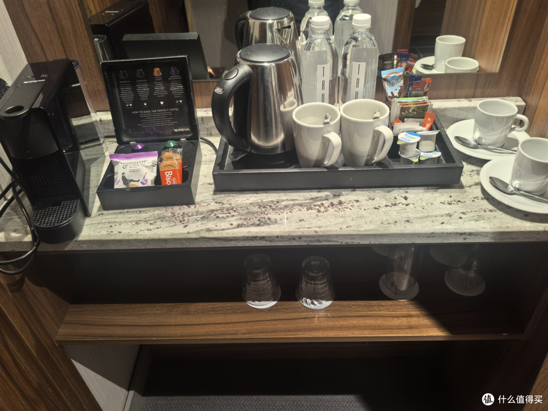 上面放着一台胶囊咖啡机和四个胶囊，两瓶饮用水、茶包、热巧克力冲剂、三包饼干、电热水壶等