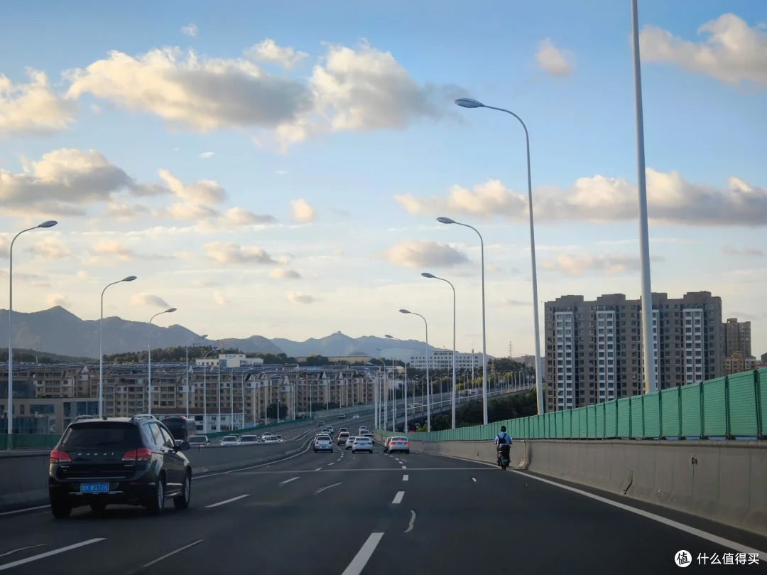 高架路上车流向前，威海市区的楼群与远山在蓝天白云下迎面铺开