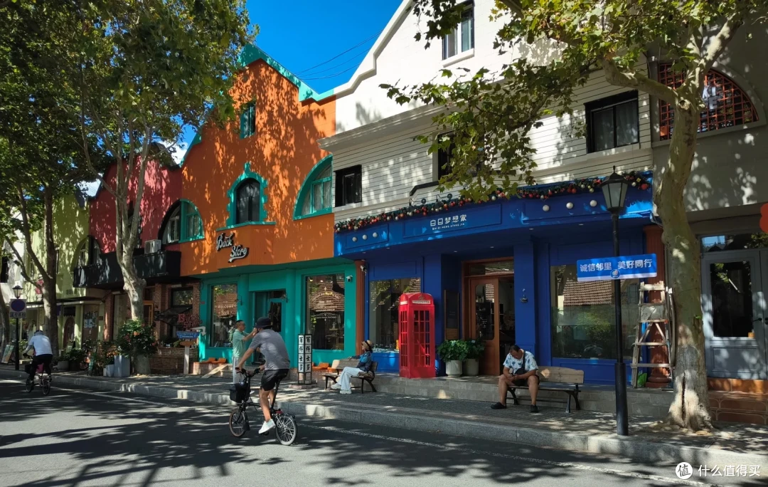 新威附路街景，橙色与蓝色的彩色小屋沿街排开，门口立着红色电话亭，梧桐树影洒满路面