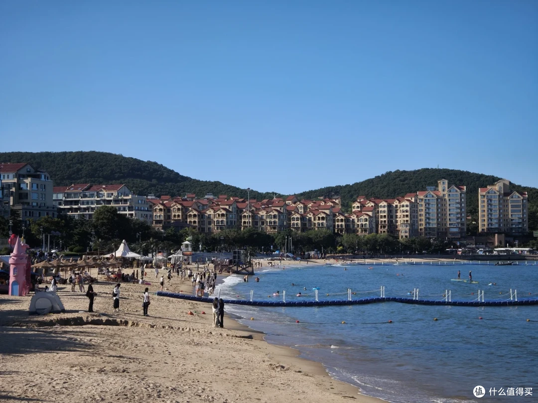 半月湾沙滩上游人戏水，蓝色浮筒围出泳区，岸后红顶楼群依山而建