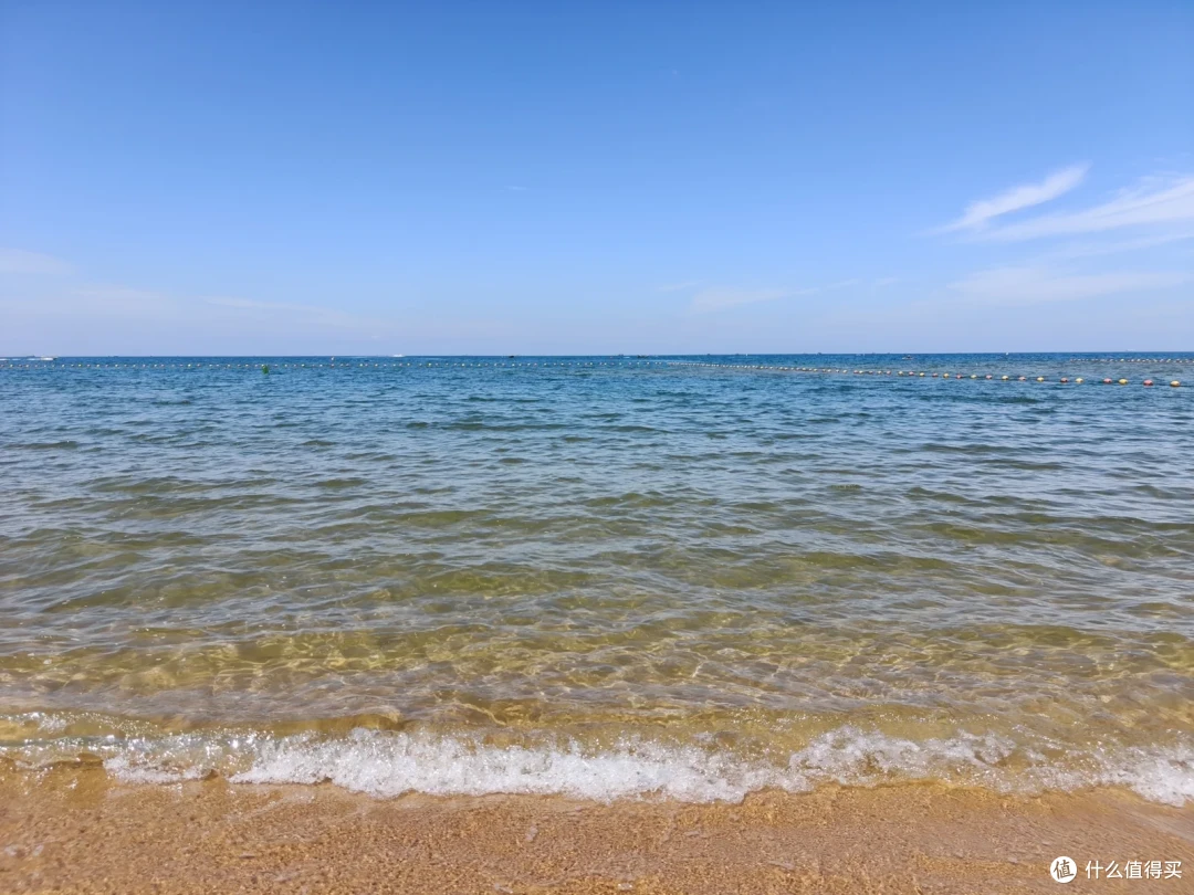 国际海水浴场清澈见底的海水泛着细浪，彩色浮球标线横贯海面，天上飘着卷云