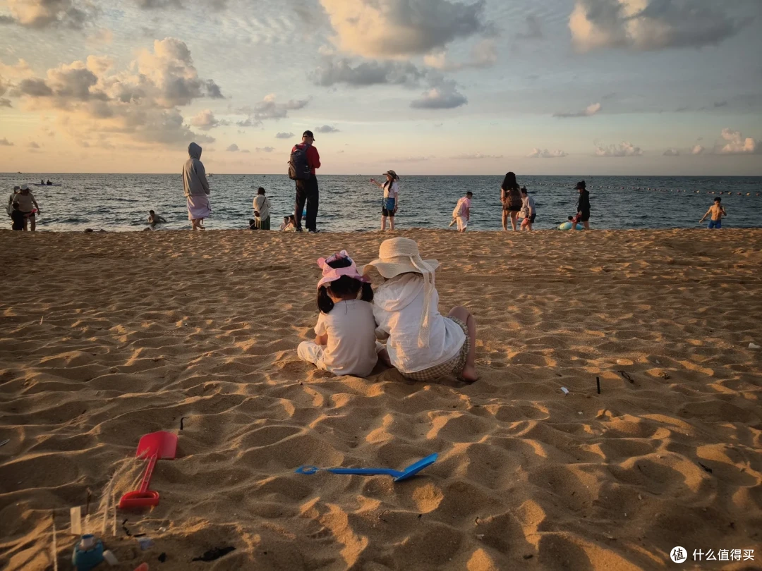 傍晚的国际海水浴场，两个戴帽子的小女孩背对镜头坐在沙滩上，身旁插着红蓝玩具铲