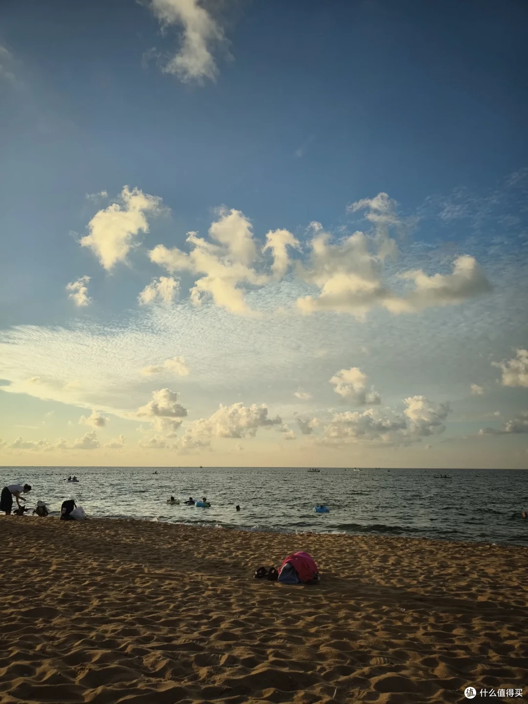 傍晚的国际海水浴场，逆光下粗颗粒沙滩上放着红背包，几个孩子在海里抱着游泳圈玩水