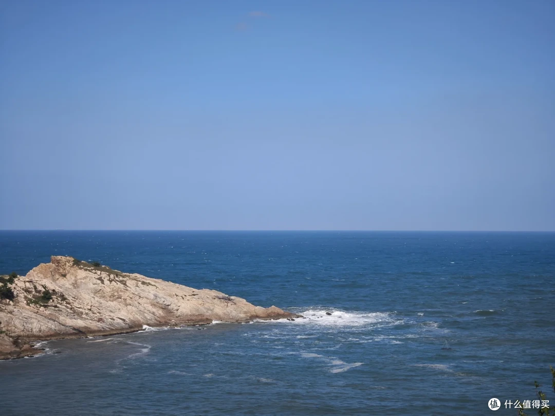 沿海公路旁的褐色礁石岬角伸入蓝色海面，浪花在岸边翻出白沫