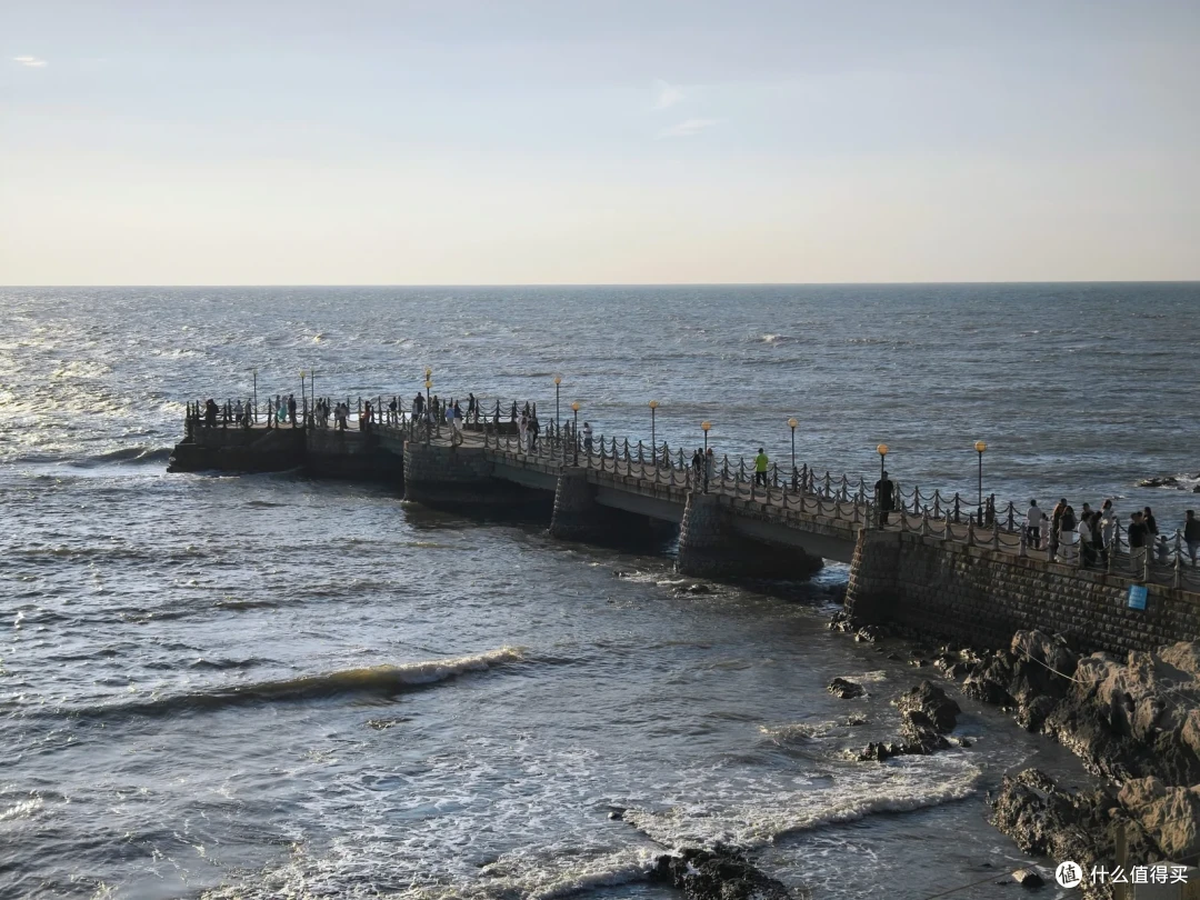 金海岸栈桥伸向波光粼粼的海面，石墩拱桥上站满游客