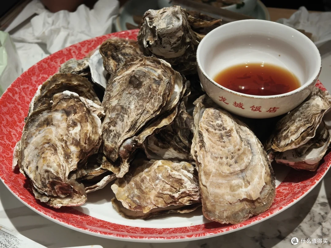 龙城饭店的一盘带壳生蚝堆在红边盘里，配一碗红色蘸料，碗沿印着店名