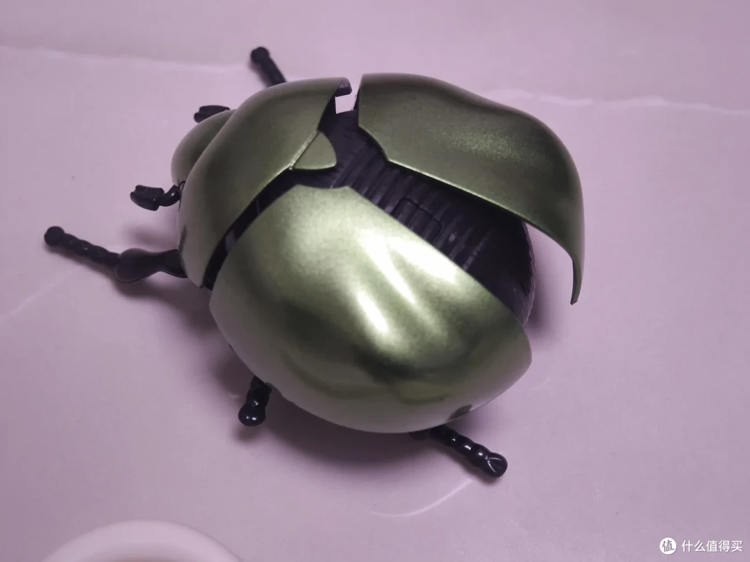 绿色金属玩具甲虫被拆开鞘翅，露出内部黑色机身和六条腿，放在浅色桌面上待修