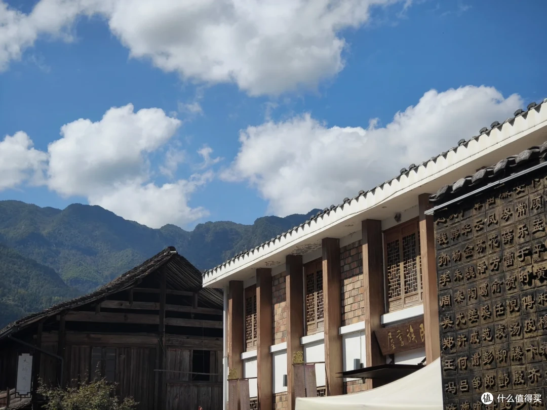 白墙青瓦的两层建筑与旁边深色木板老屋，右墙刻满汉字姓氏，背后是连绵青山