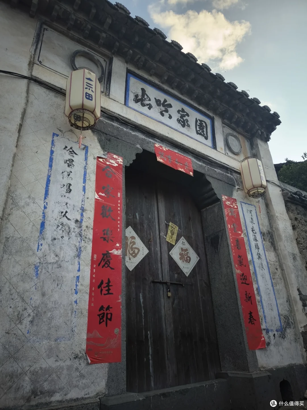 白墙老宅的深色木门前贴着红春联与褪色的蓝字旧联，门额题着山哈家园