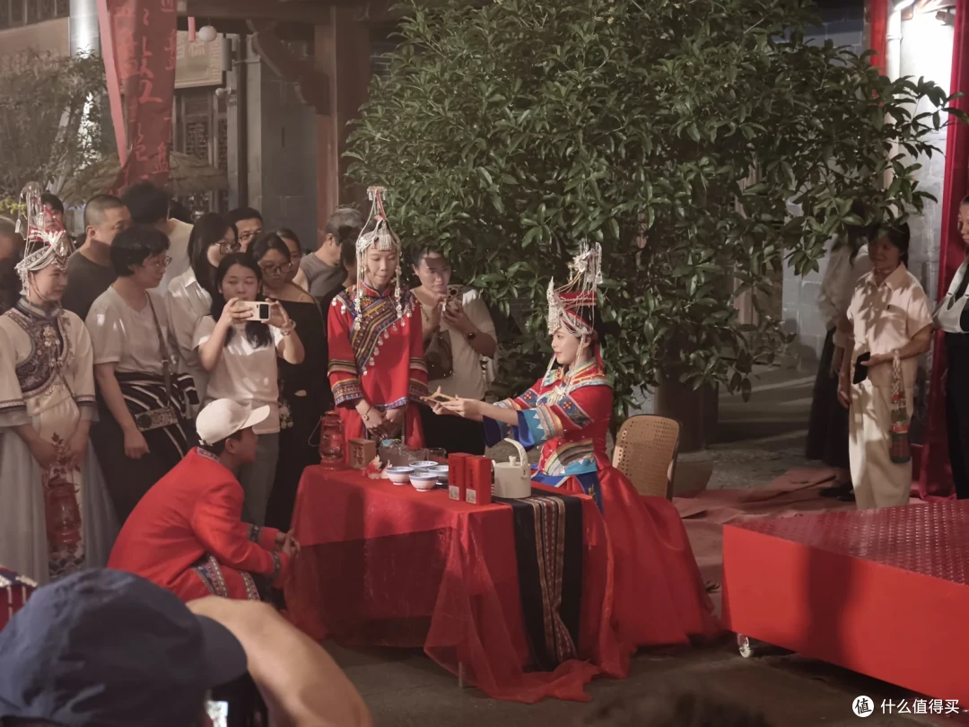 铺红布的桌前，穿红色婚服的女子向跪着的男子敬茶，旁有人举手机拍摄