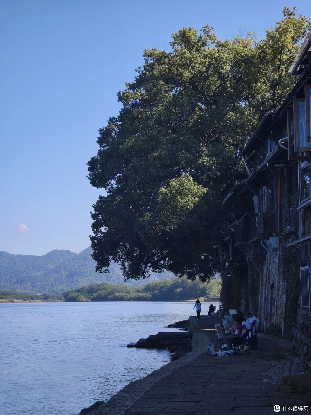 江边步道旁的大树遮出浓荫，几个人坐在岸边石阶上