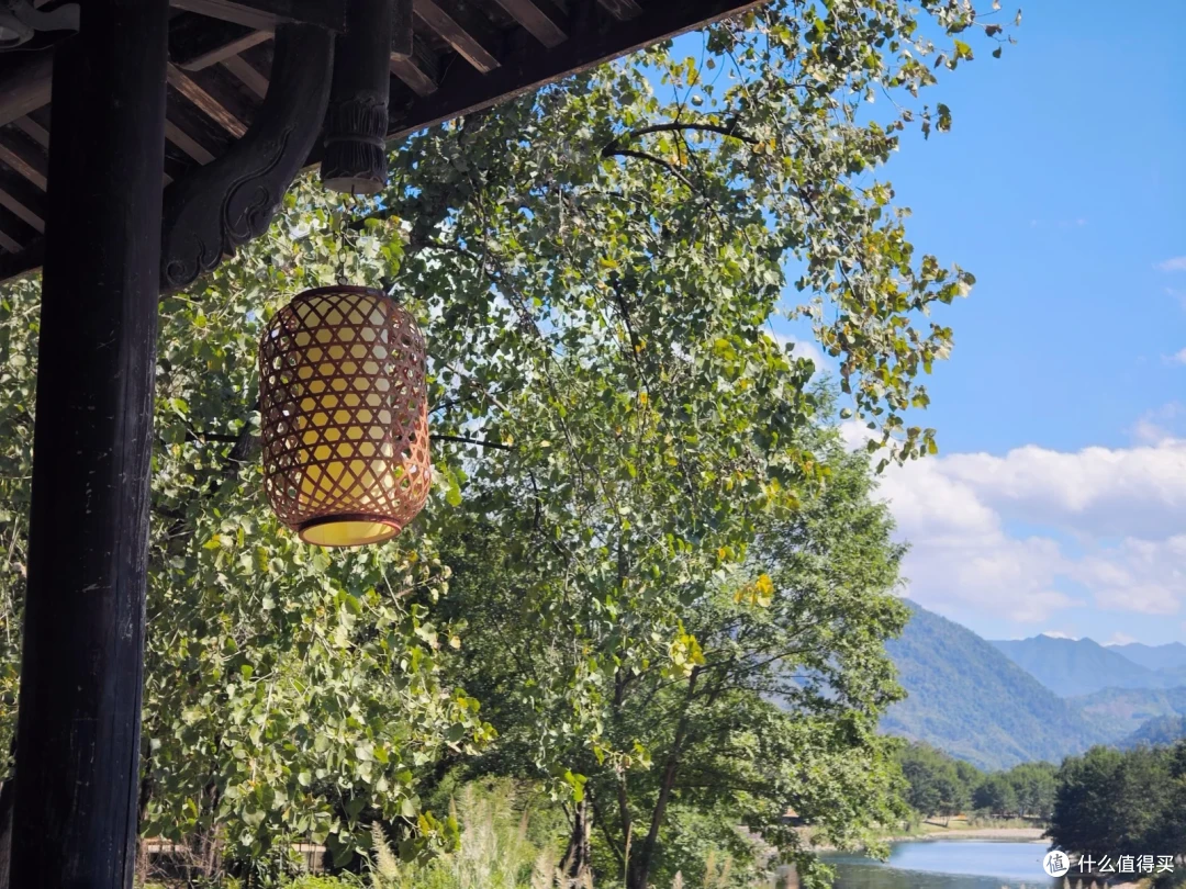 屋檐下垂着一只竹编灯笼，前景是树叶，远处是江面与青山