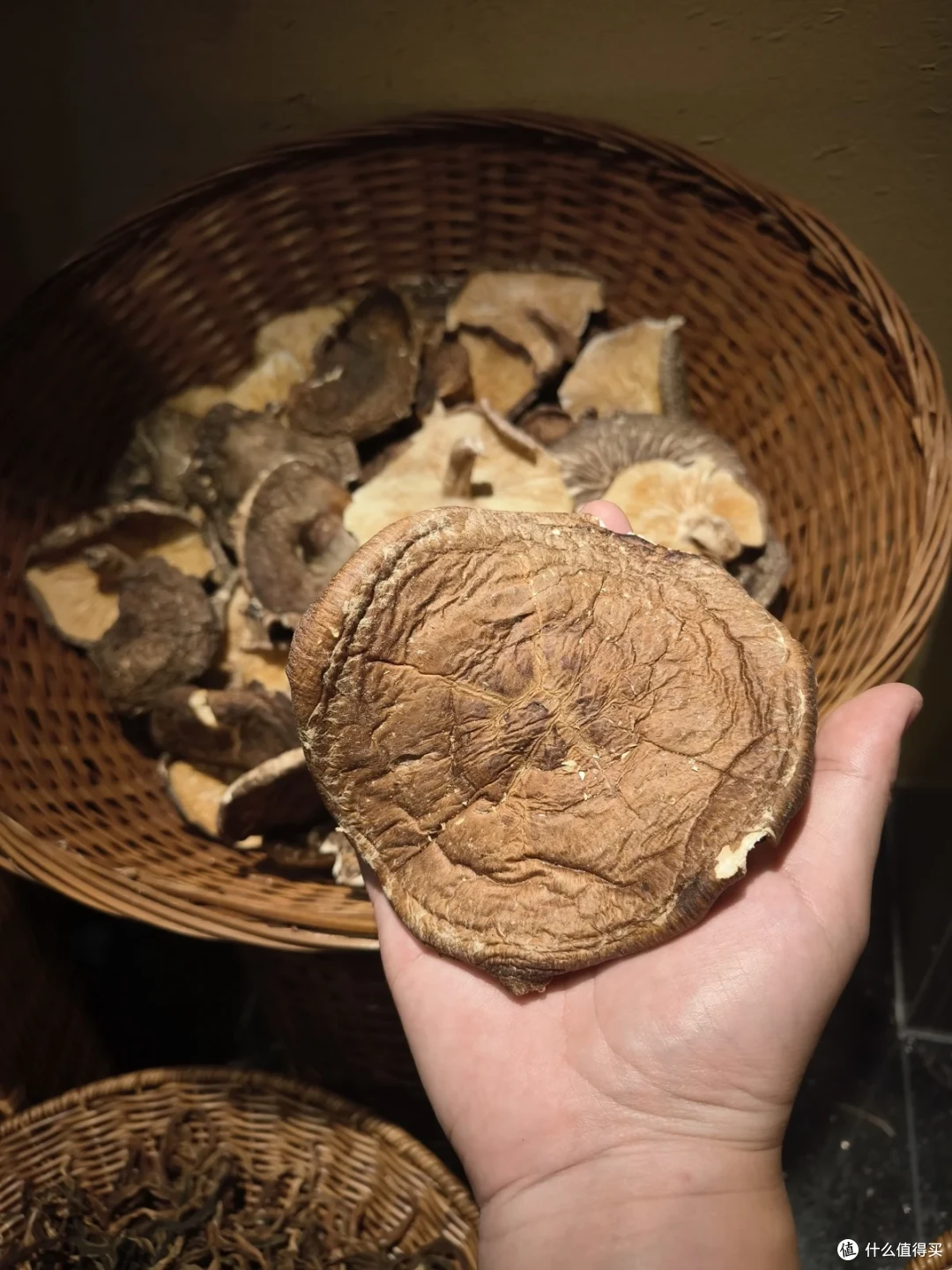 一只手举着一朵巴掌大的干香菇，下方是一筐同样的干货